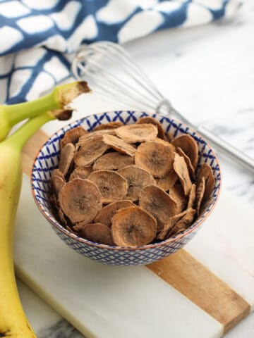 A small bowl of cocoa-dusted banana chips on a marble board next to two whole bananas