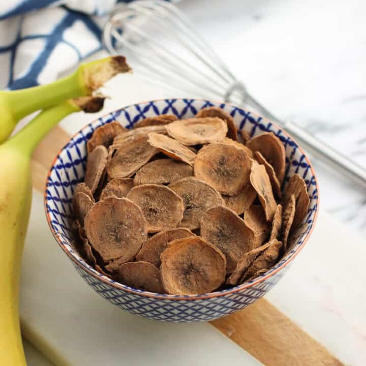 A small bowl of cocoa-dusted banana chips on a marble board next to two whole bananas