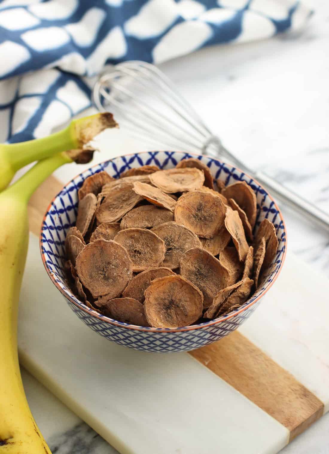 A small ceramic bowl filled with banana chips next to fresh bananas and a small whisk