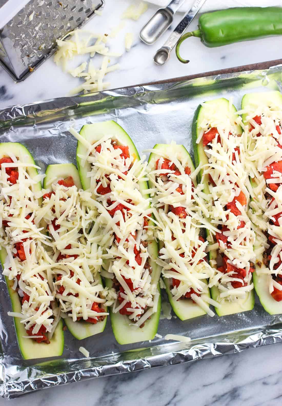 Hollowed out zucchini halves on a foil-lined baking sheet topped with beef, tomatoes, and shredded cheese