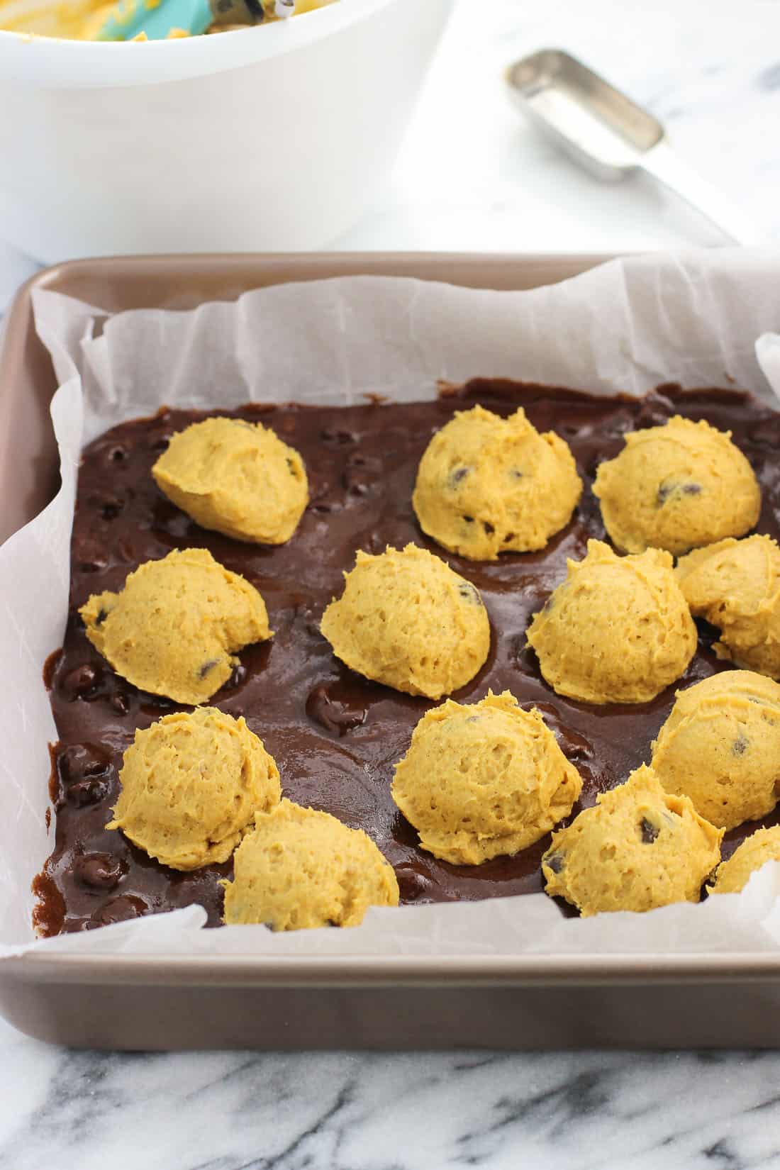 Brownie batter in a parchment-lined baking dish with globs of pumpkin batter scattered on top before being swirled in.
