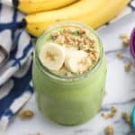 A smoothie in a mason jar topped with banana slices and granola with a bunch of bananas in the background