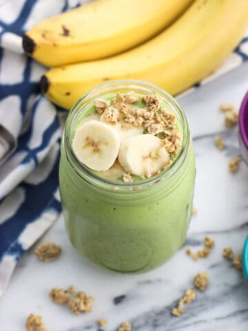 A smoothie in a mason jar topped with banana slices and granola with a bunch of bananas in the background