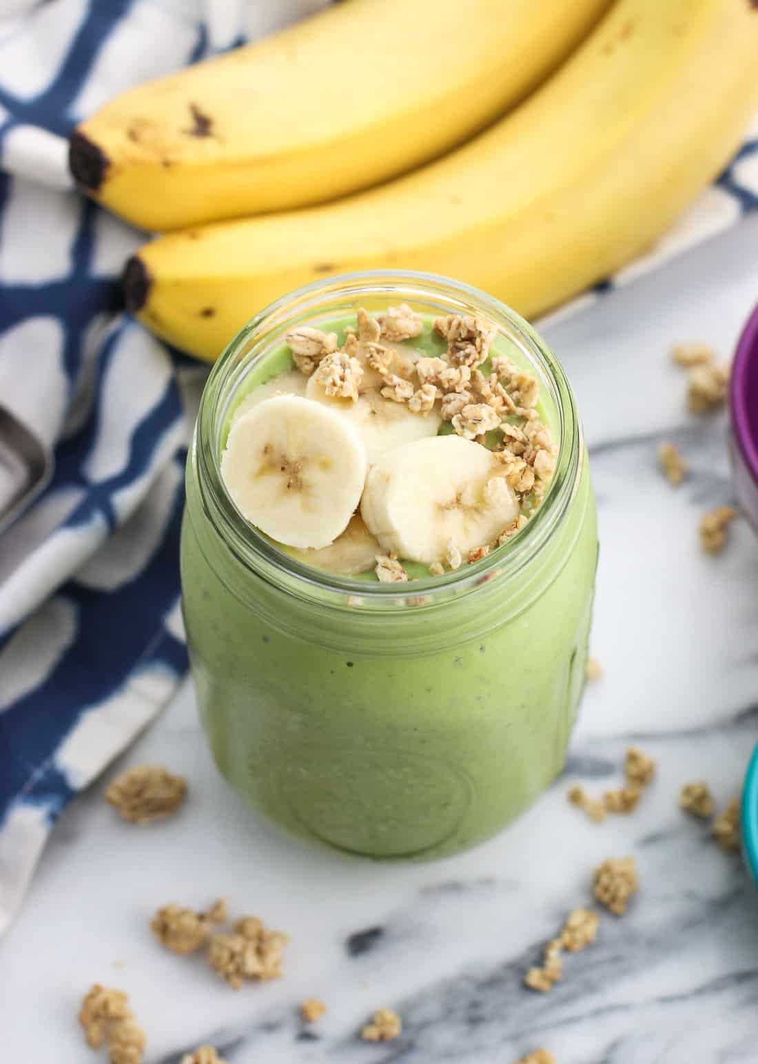 A smoothie in a mason jar topped with banana slices and granola with a bunch of bananas in the background