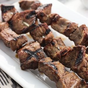 A close-up of three parallel stainless steel skewers of grilled steak on a ceramic tray