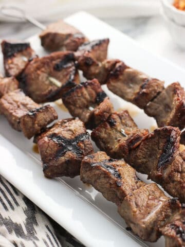 A close-up of three parallel stainless steel skewers of grilled steak on a ceramic tray