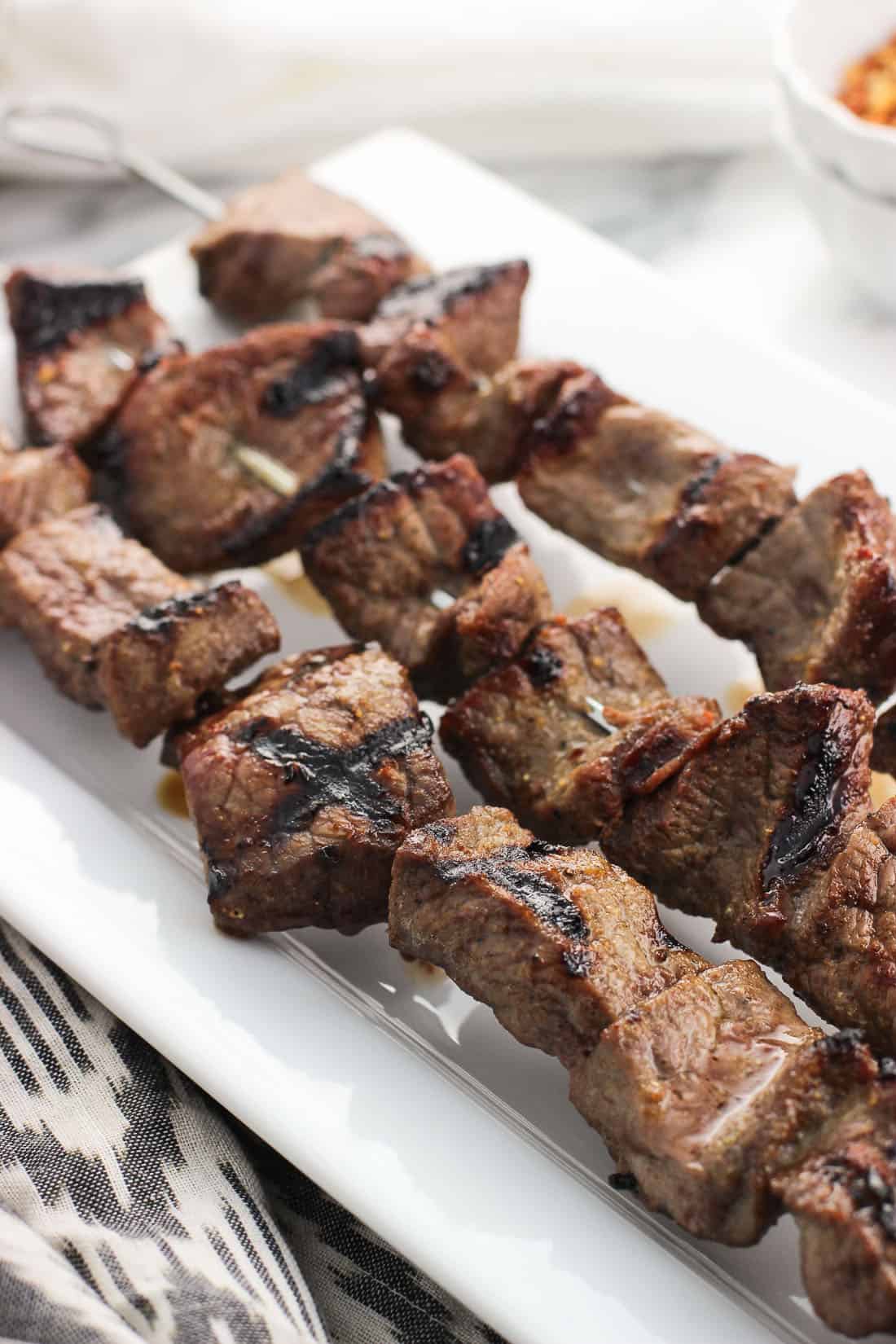 A close-up of three parallel stainless steel skewers of grilled steak on a ceramic tray