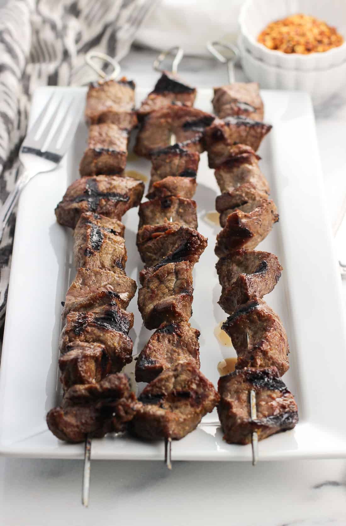 Three stainless steel skewers of steak bites on a rectangular tray with seasoning and a fork in the background