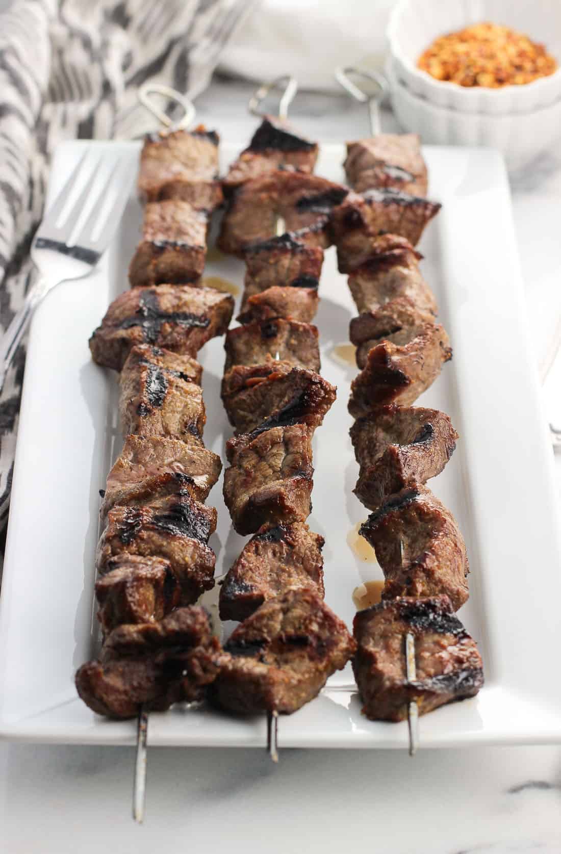 Three stainless steel skewers of steak bites on a rectangular tray with seasoning and a fork in the background.