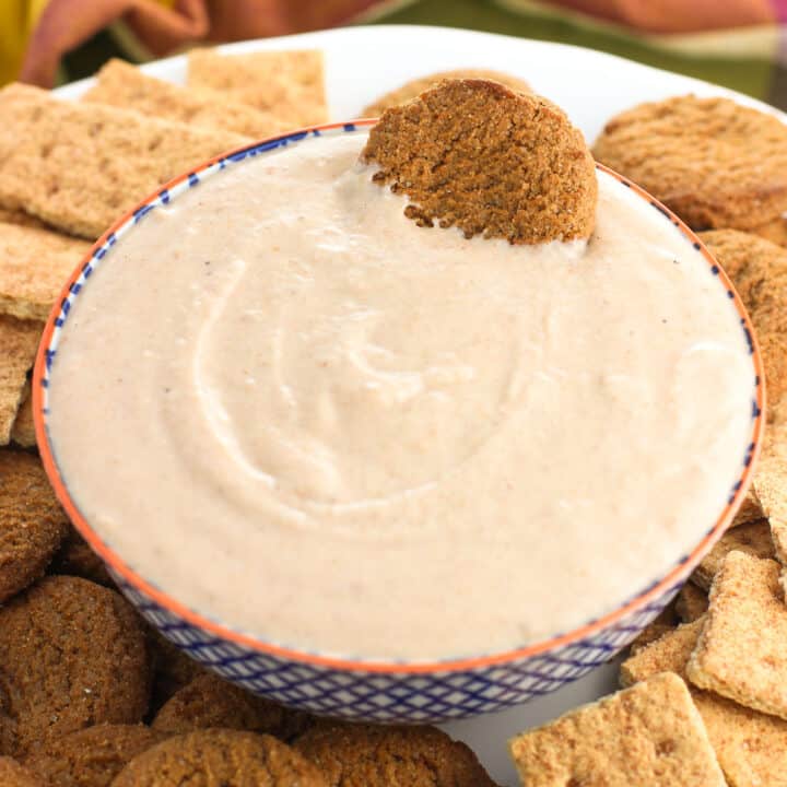 A bowl of yogurt dip on a plate with graham crackers and gingersnap cookies with a gingersnap dunked in