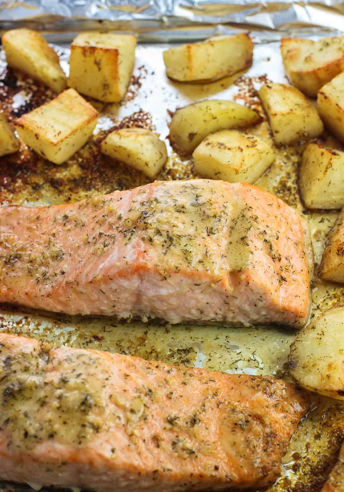 Two cooked salmon fillets coated in horseradish sauce on a foil-lined baking sheet surrounded by potato wedges