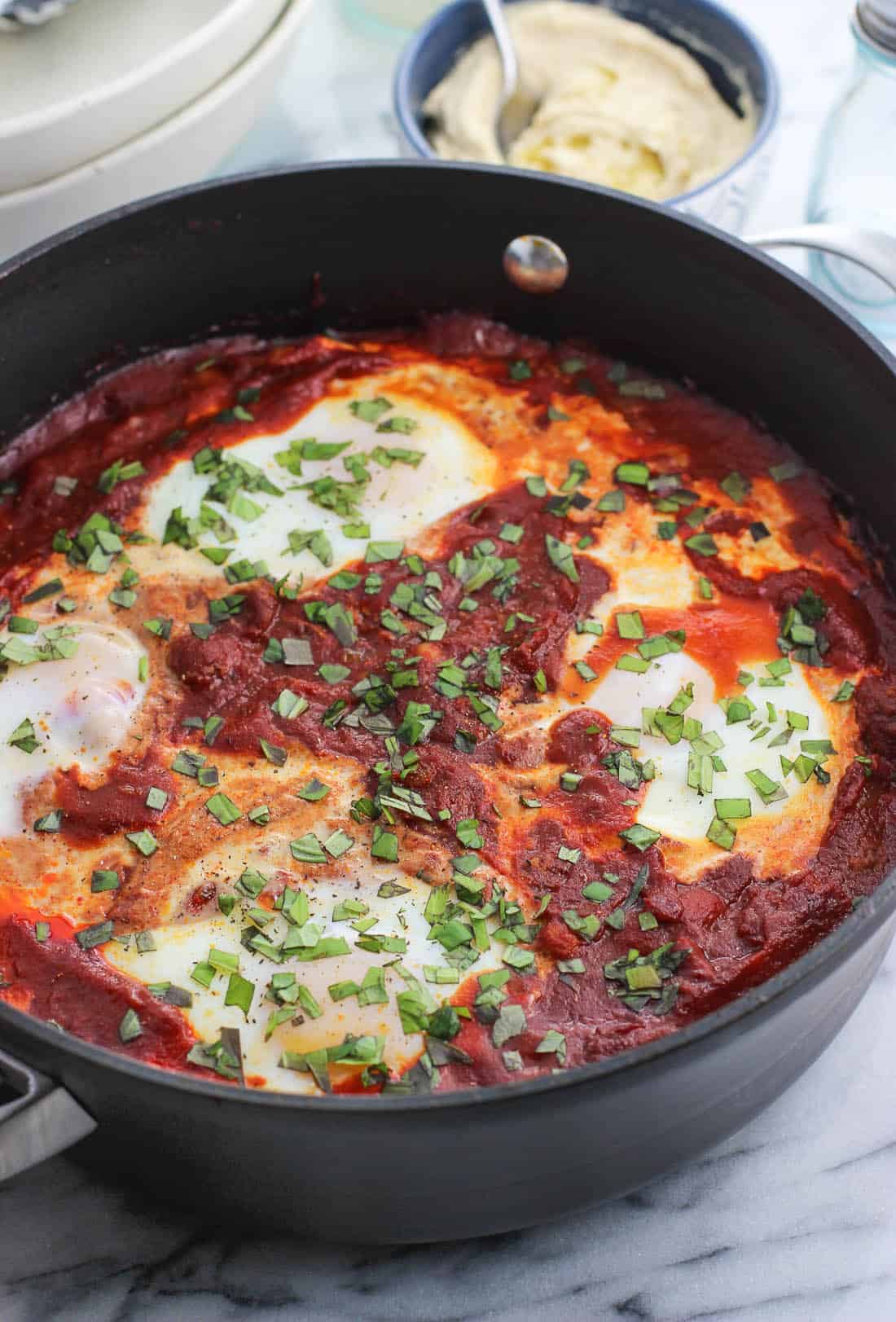 Shakshuka with four eggs in a tall-sided skillet topped with herbs with a small bowl of hummus in the background
