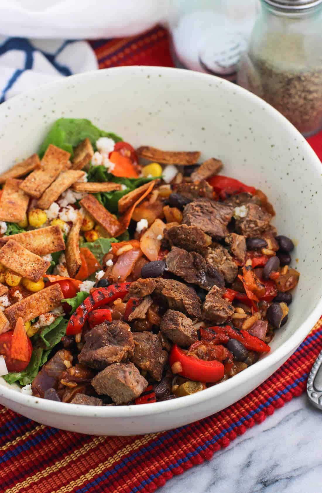 Barbacoa beef and peppers in a bowl next to a salad with salt and pepper shakers in the background.