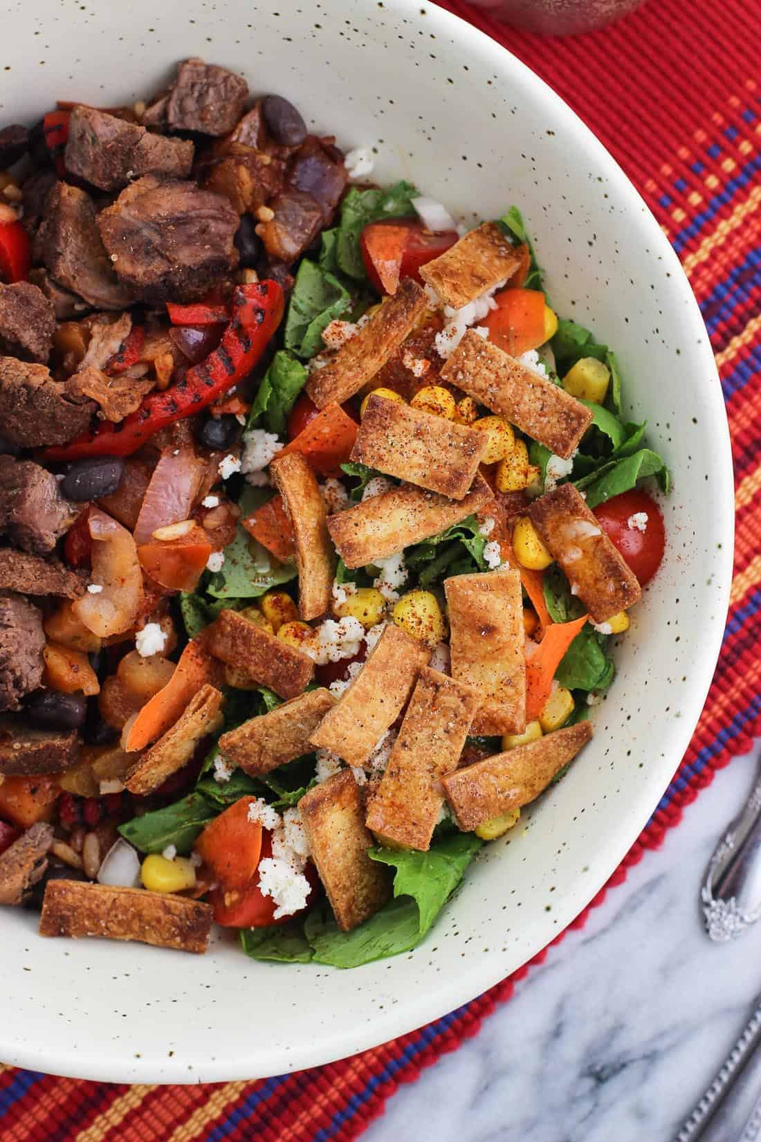 A side salad topped with tortilla strips in a bowl with steak and peppers.