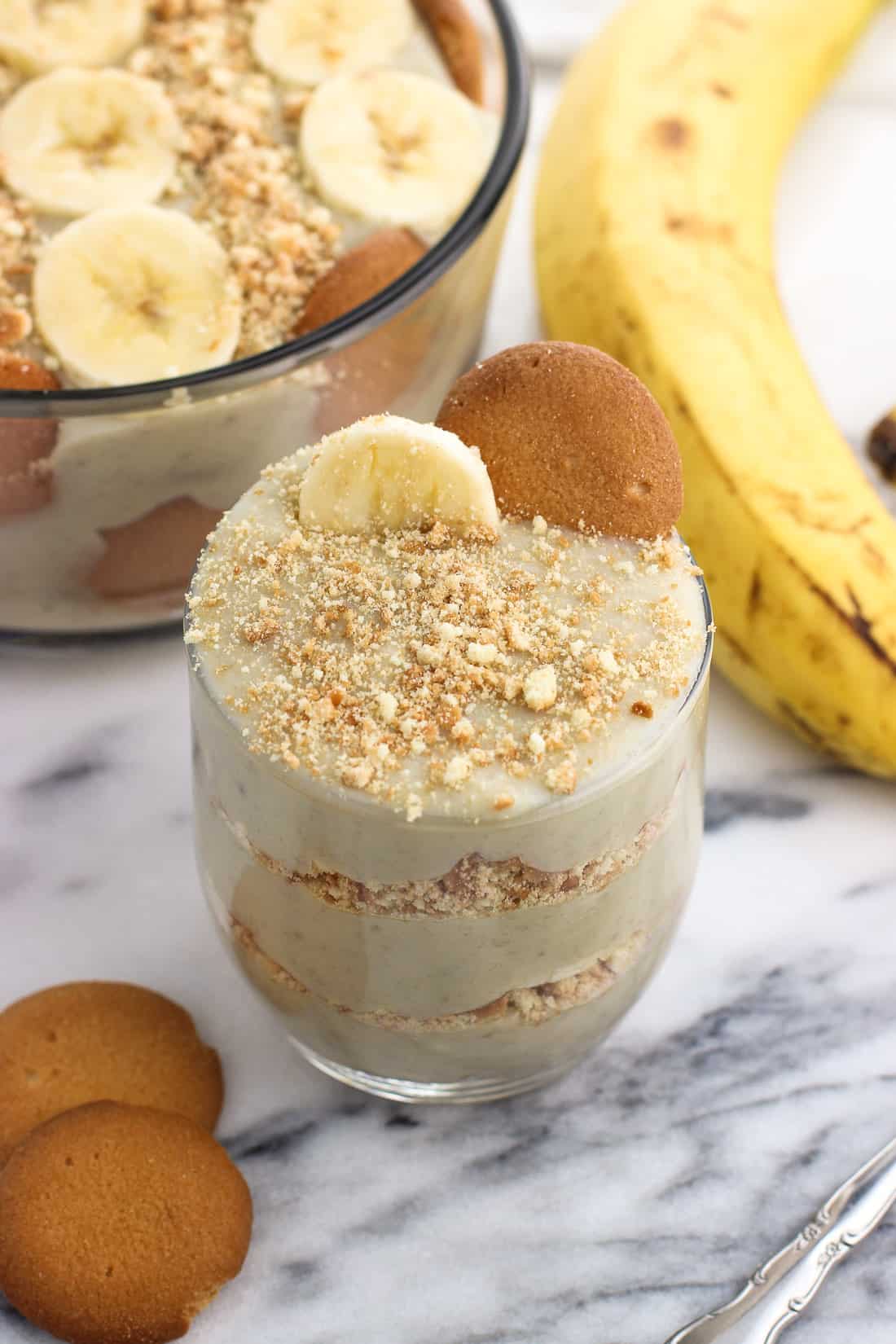 A small cup of layered banana pudding next to wafers and a banana.