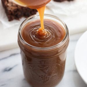 Caramel sauce being poured into a glass mason jar with a batch of brownies on a board in the background