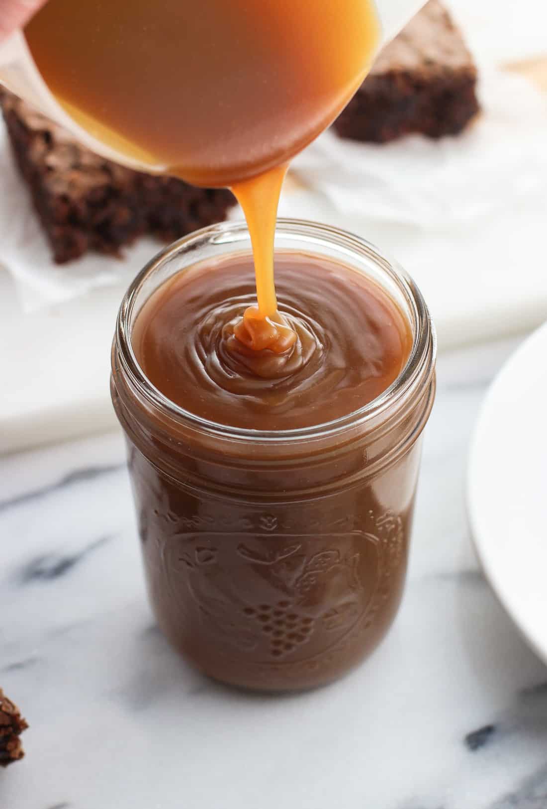 Caramel sauce being poured into a glass mason jar with a batch of brownies on a board in the background.