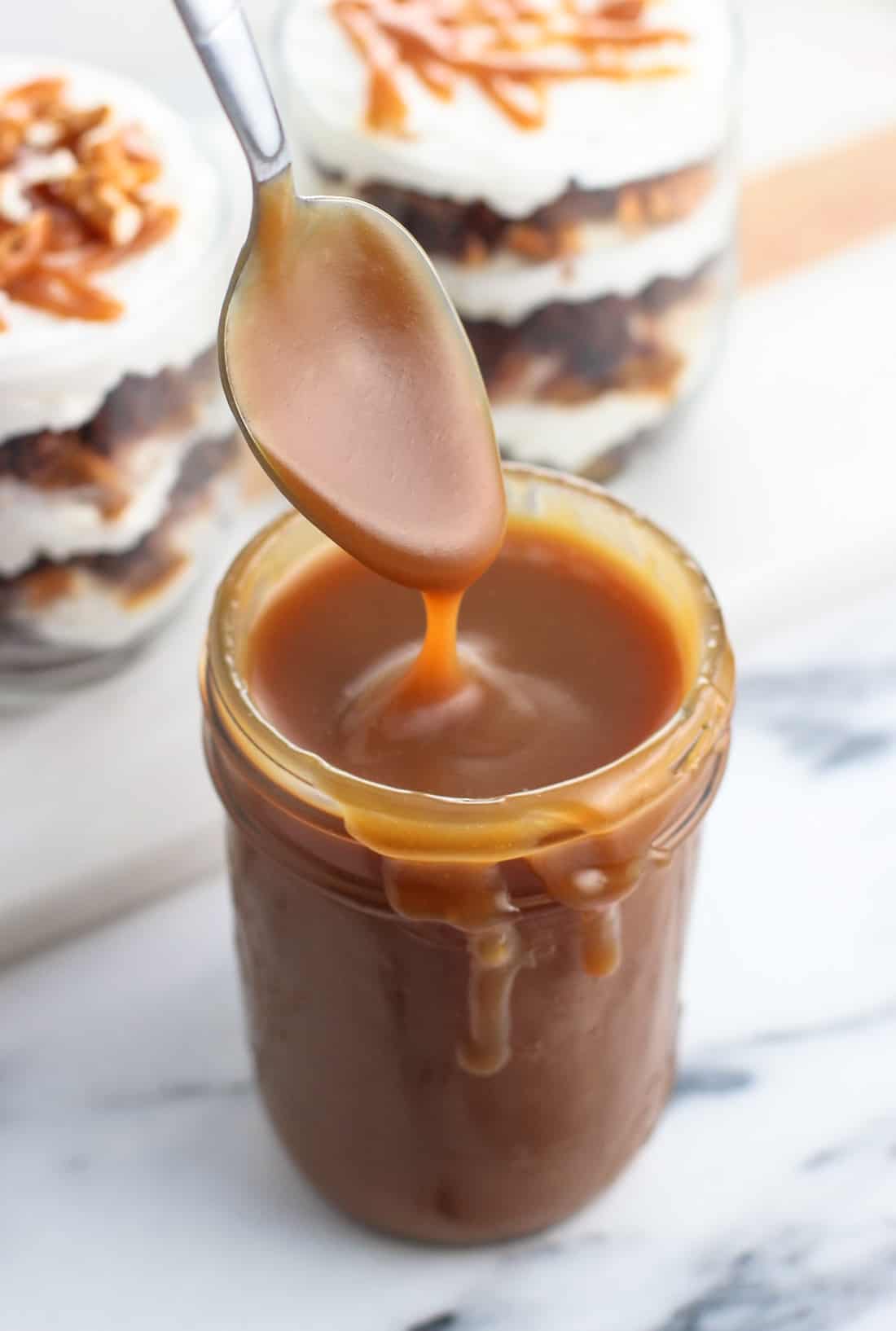 A spoon being dipped in a jar of caramel sauce with two mini brownie parfaits in the background