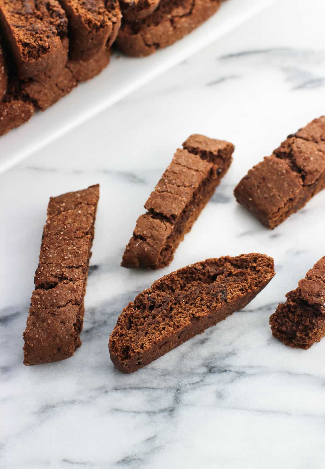 Five biscotti on a marble board in front of a full tray of them