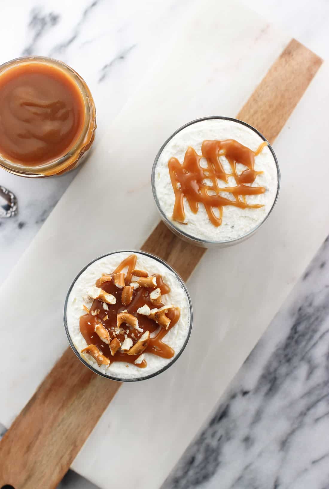 An overhead picture of a jar of caramel sauce and two individual trifle servings on a marble board, one topped with crushed pretzels