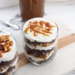 Two brownie trifles in small glasses next to a jar of caramel sauce.