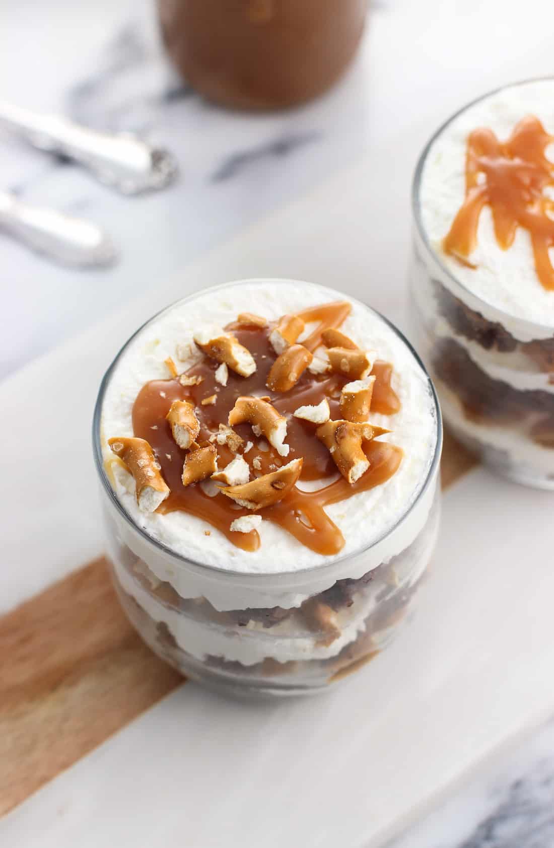 Two small glass cups filled with whipped cream, brownies, pretzels, and caramel sauce