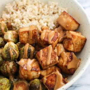 A bowl of baked tofu and roasted brussels sprouts halves drizzled with peanut sauce served with brown rice