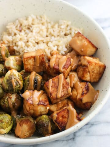 A bowl of baked tofu and roasted brussels sprouts halves drizzled with peanut sauce served with brown rice