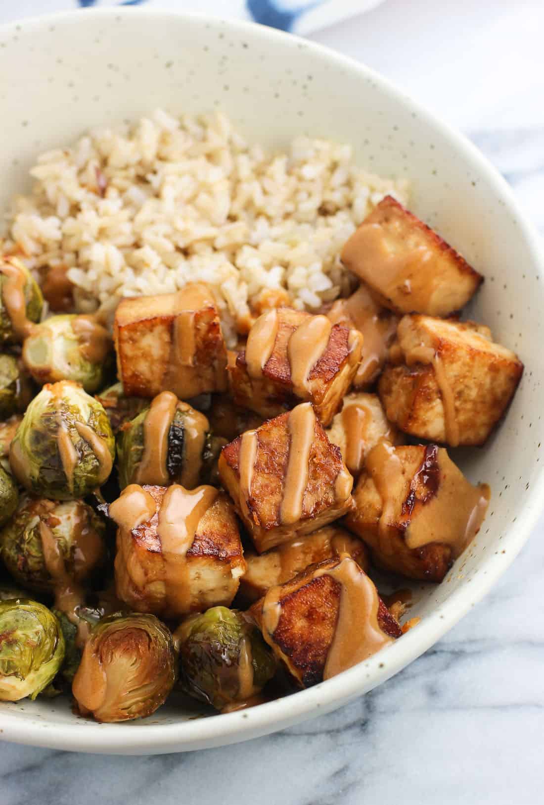 A bowl of baked tofu and roasted brussels sprouts halves drizzled with peanut sauce served with brown rice