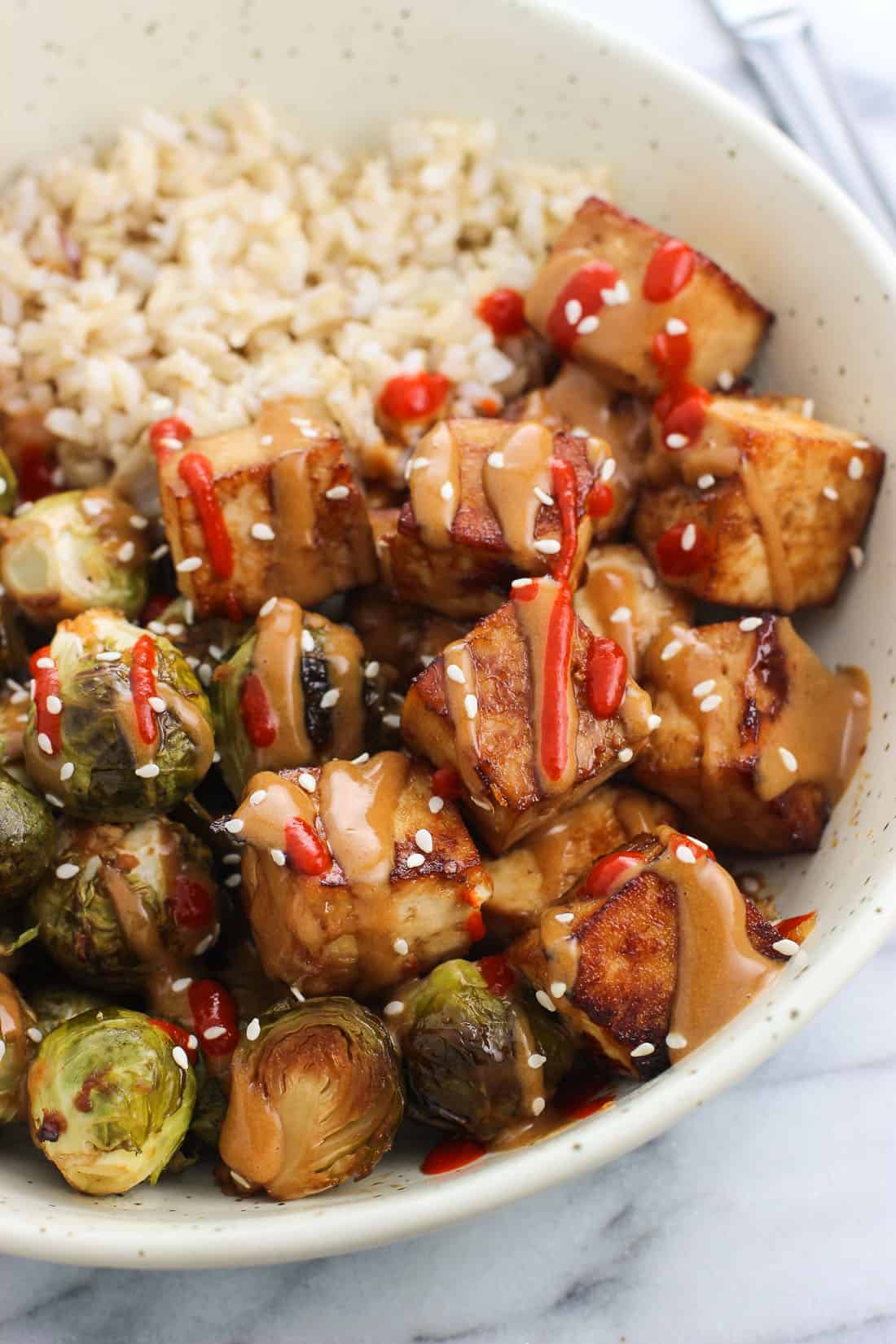 A close-up of roasted tofu cubes and brussels sprouts drizzled with peanut sauce and sriracha, served next to brown rice.