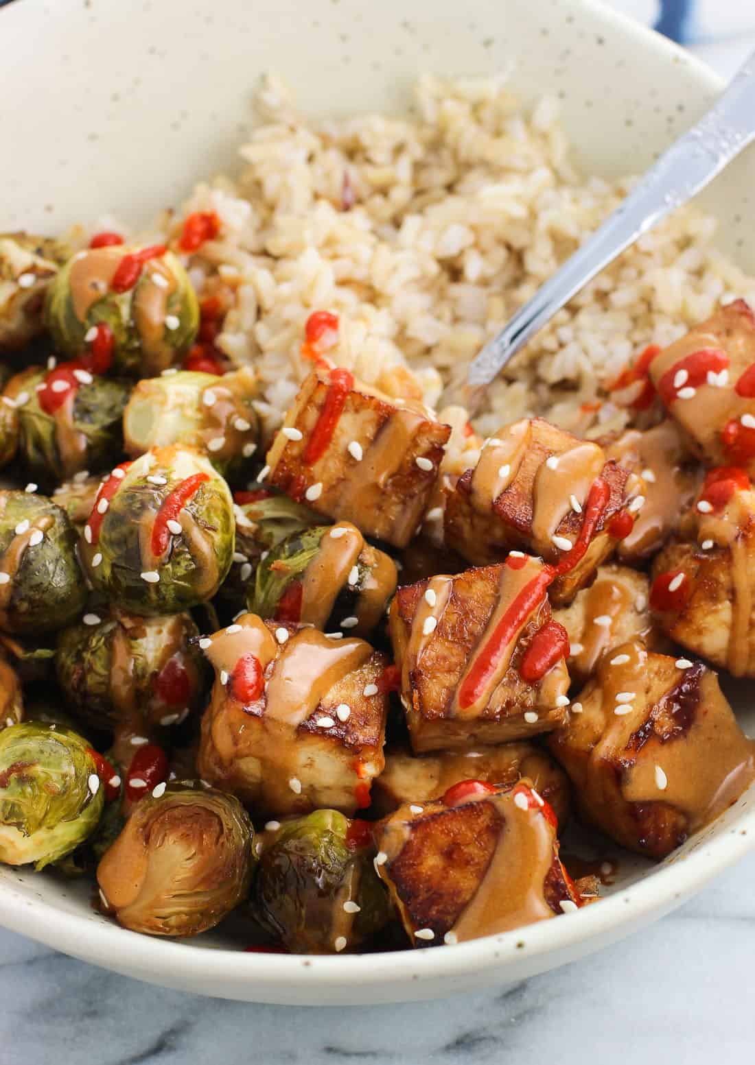 The finished dish (roasted tofu, brussels sprouts, rice) in a bowl drizzled with peanut sauce, sriracha, and sesame seeds with a fork.