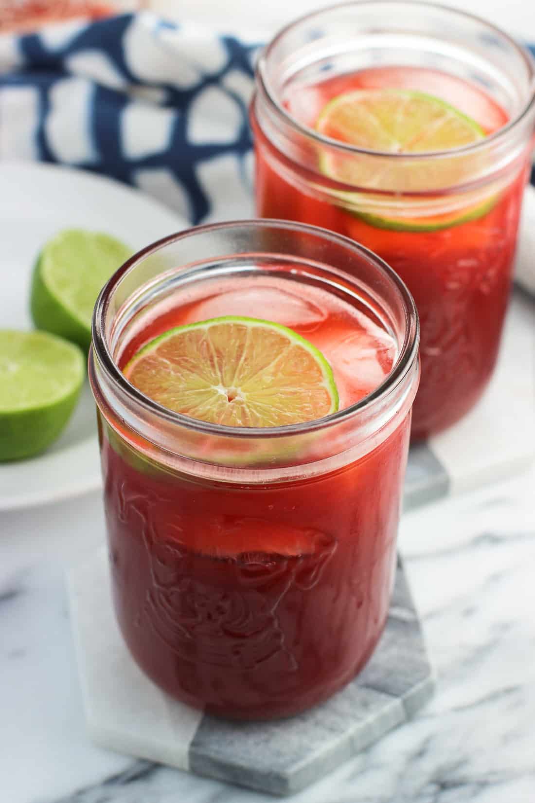 Two glasses of strawberry drinking vinegar on marble coasters next to a dish towel and lime rounds