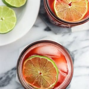 An overhead view of two glasses of drinking vinegar garnished with lime rounds and a plate of lime halves