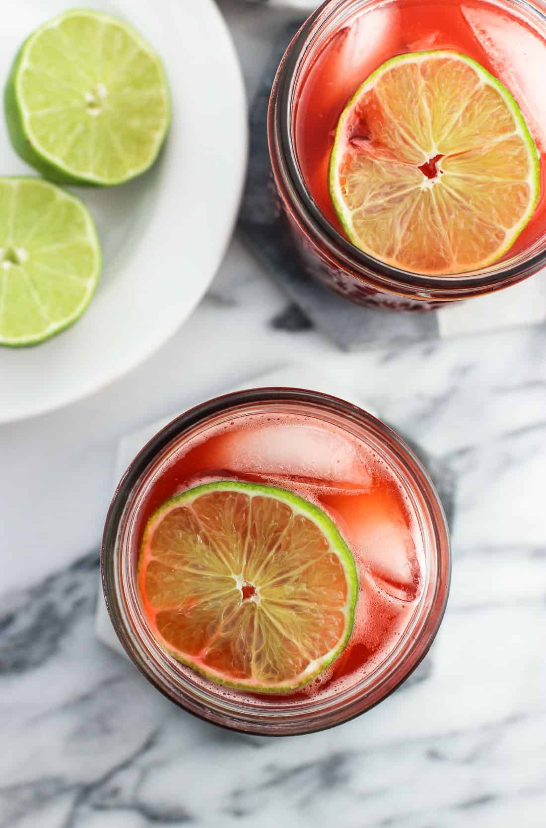 An overhead shot of two glasses of drinking vinegar garnished with a lime round next to lime halves on a plate