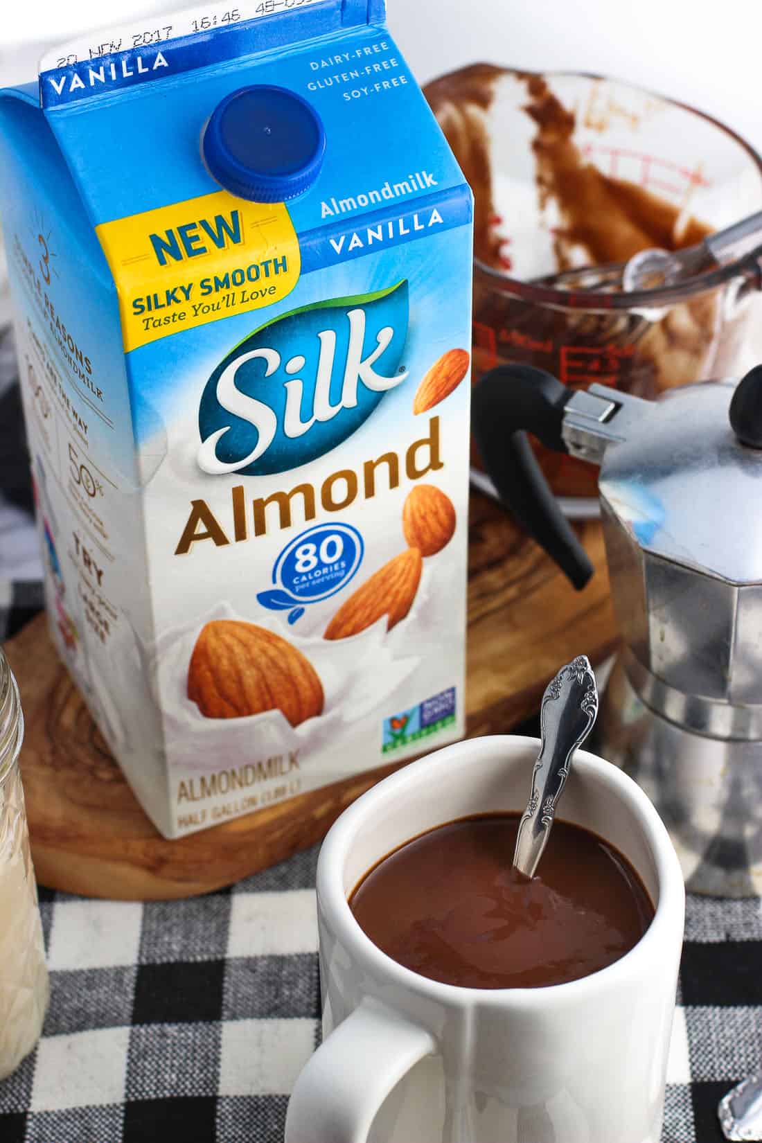 A mug of mocha hot chocolate next to a carton of almond milk and a small espresso pot.