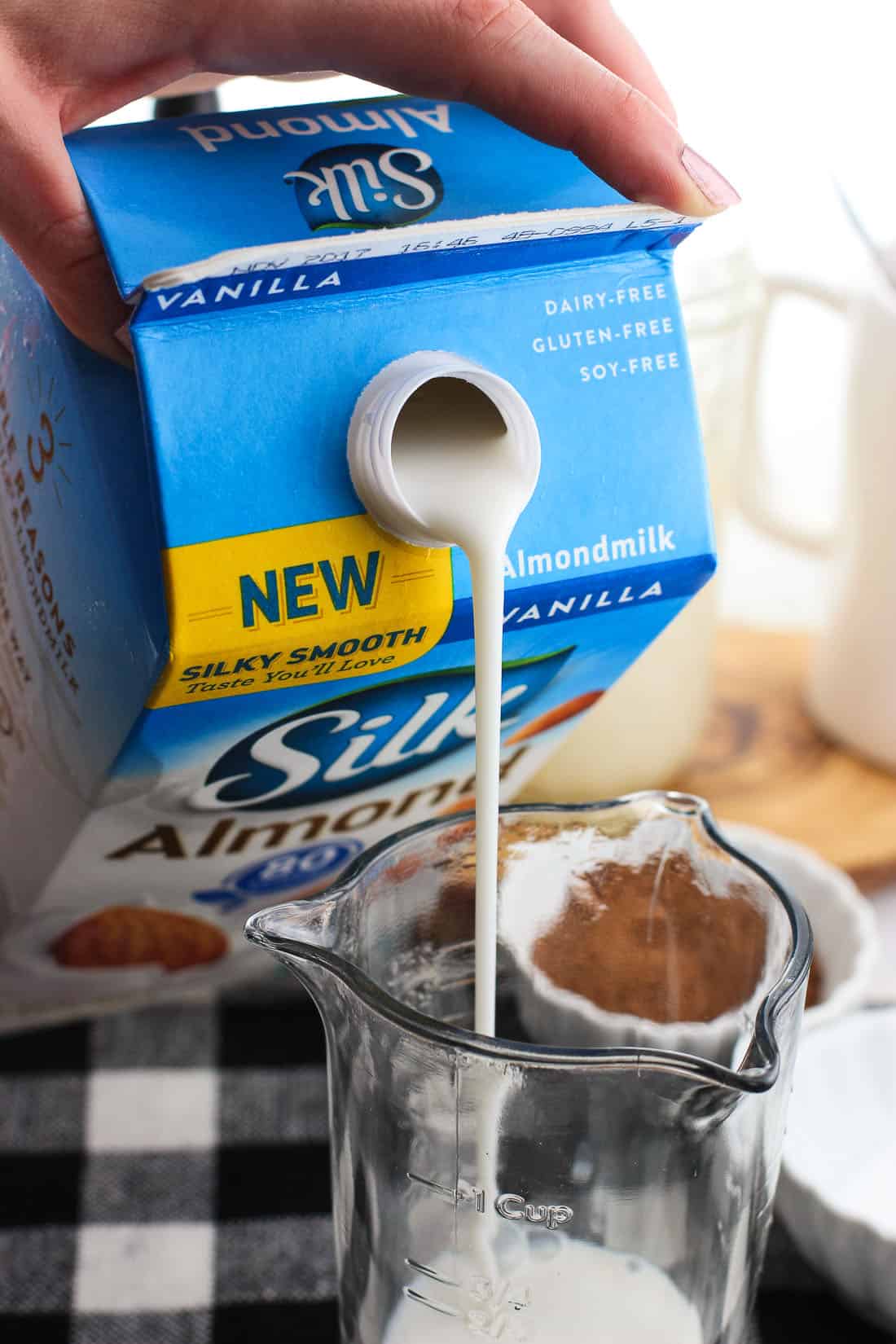 A carton of vanilla almond milk being poured into a glass measuring cup.