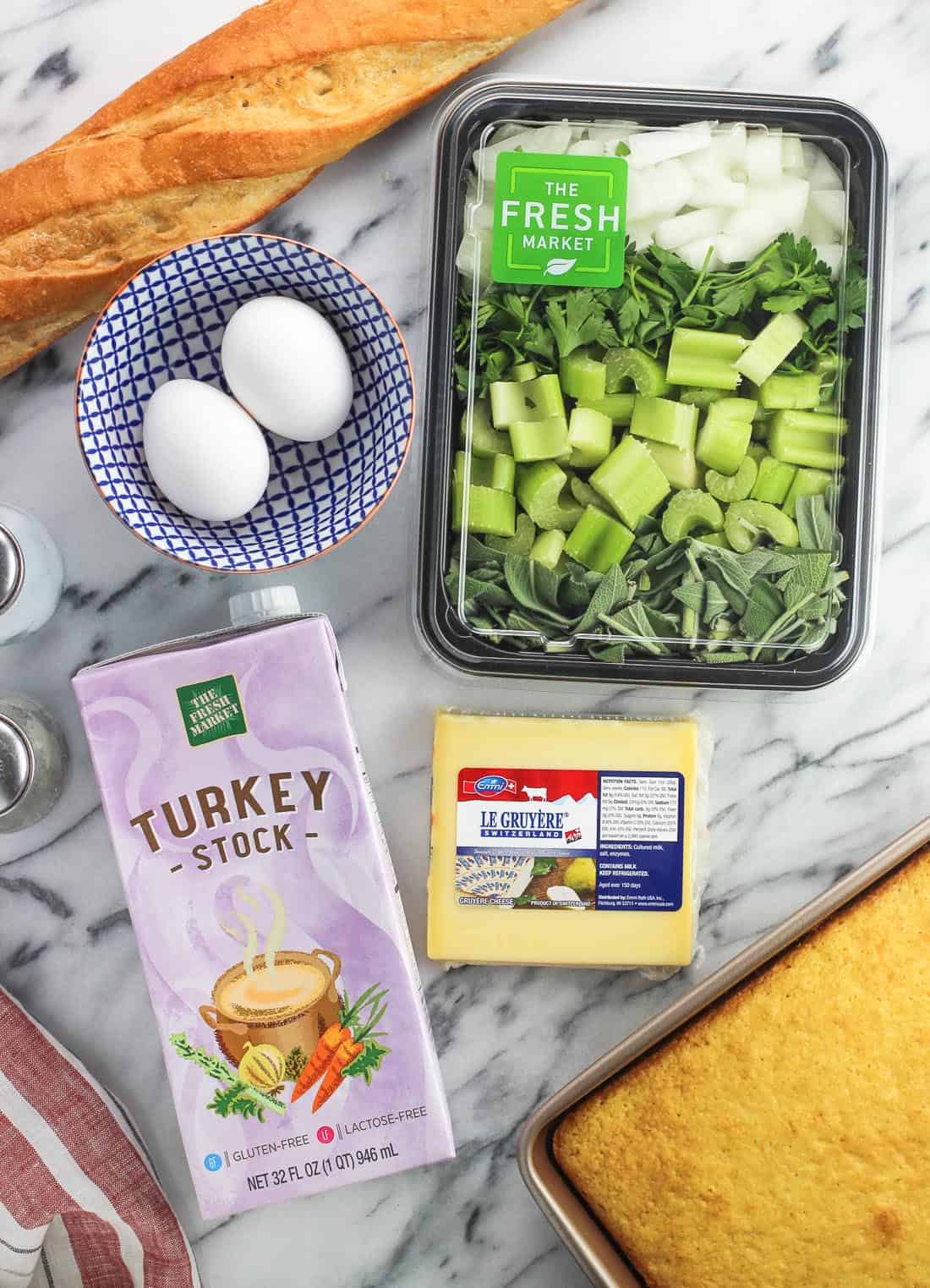 Recipe ingredients on a marble board, including a baguette, eggs, celery, herbs, onion, Gruyere, a carton of turkey stock, and a pan of cornbread