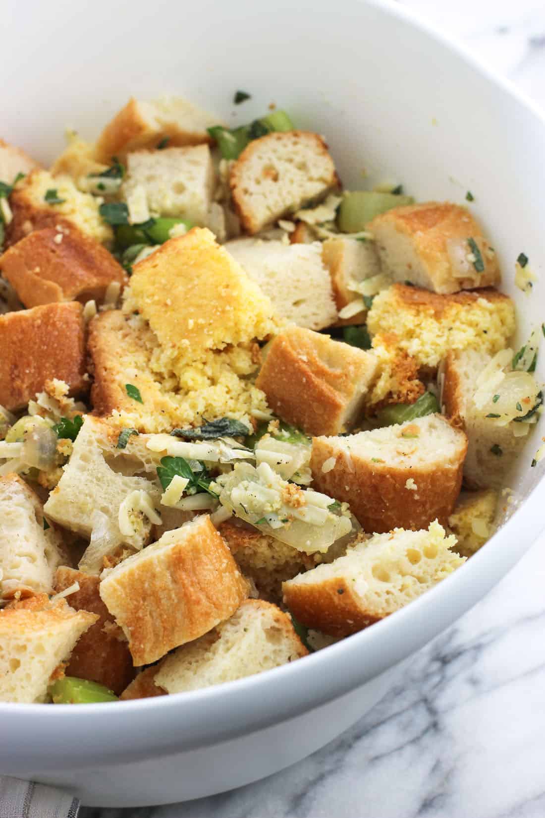 Cornbread and baguette cubes, grated cheese, diced herbs, and vegetables in a large mixing bowl