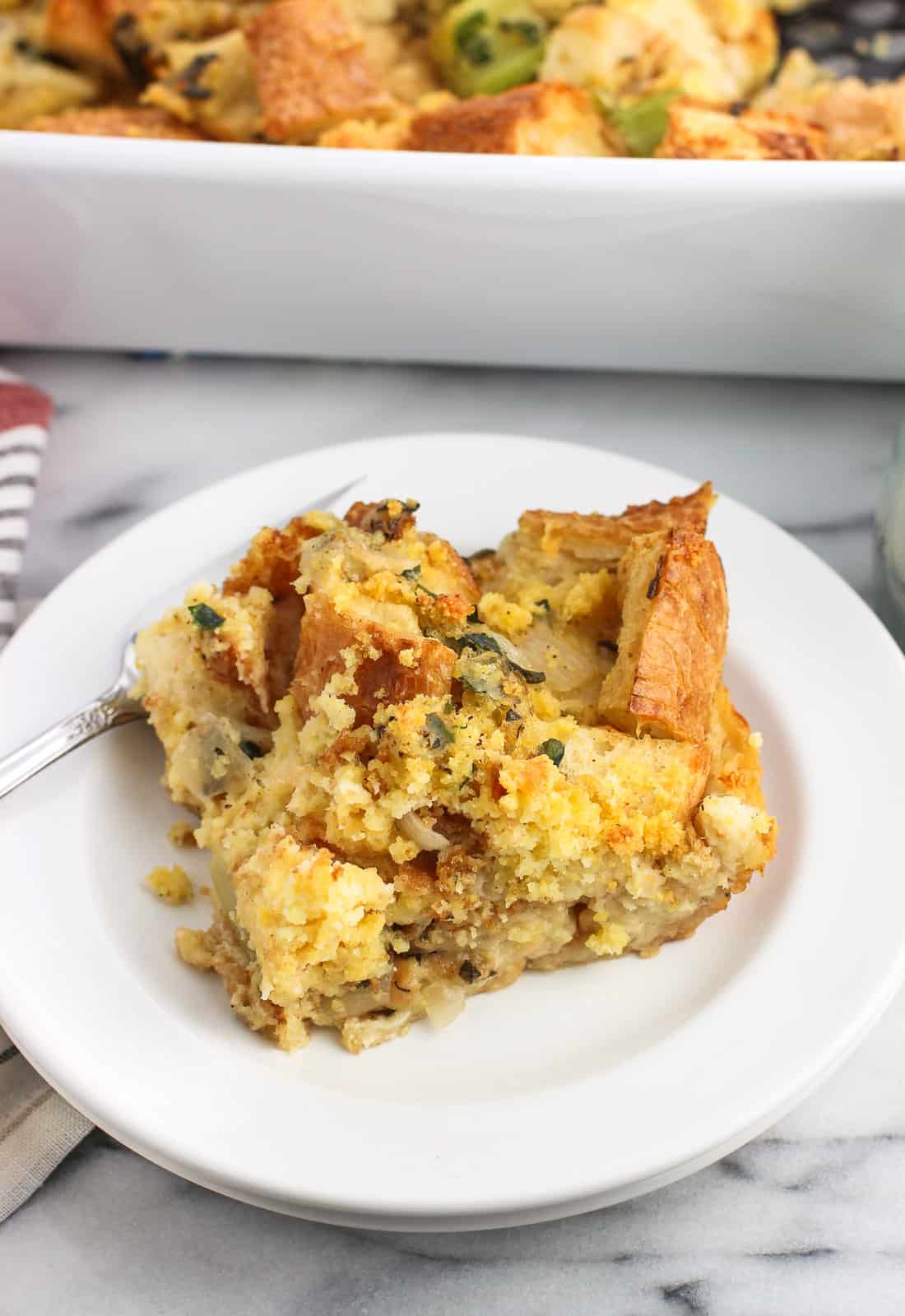 A slice of cornbread strata on a small plate with a fork with the whole pan in the background