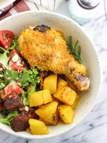 A baked chicken drumstick on a dish next to roasted potatoes and a side salad including tomatoes and croutons