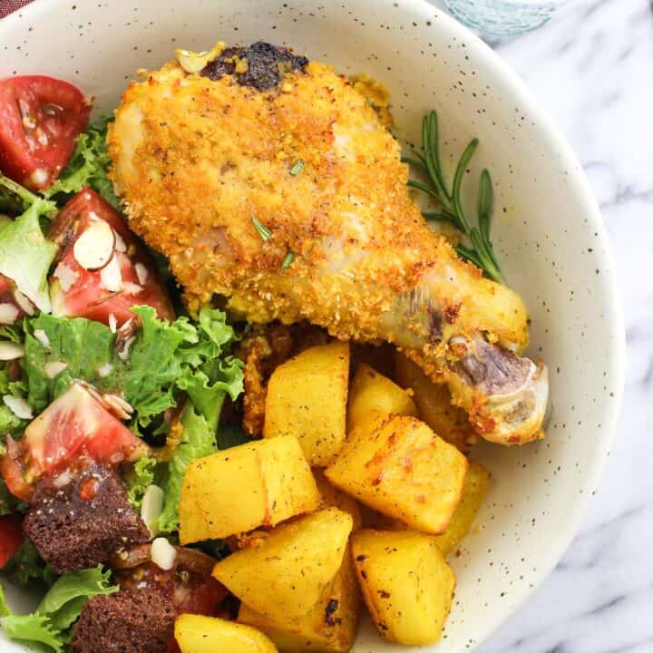 A baked chicken drumstick on a dish next to roasted potatoes and a side salad including tomatoes and croutons