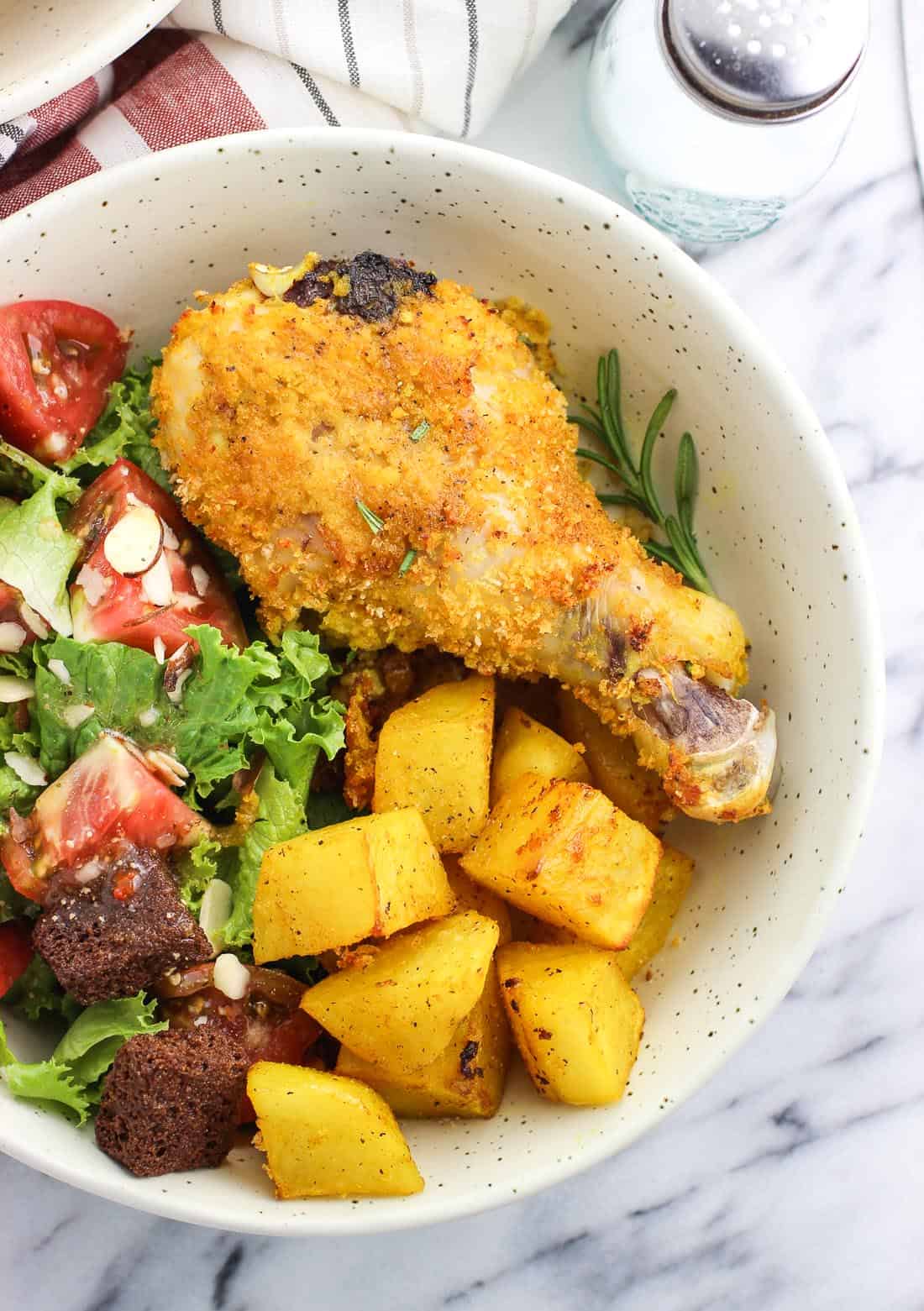 A baked chicken drumstick on a dish next to roasted potatoes and a side salad including tomatoes and croutons