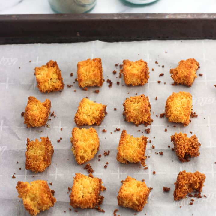 Sixteen cornbread croutons on a parchment-lined baking sheet with salt and pepper shakers in the background
