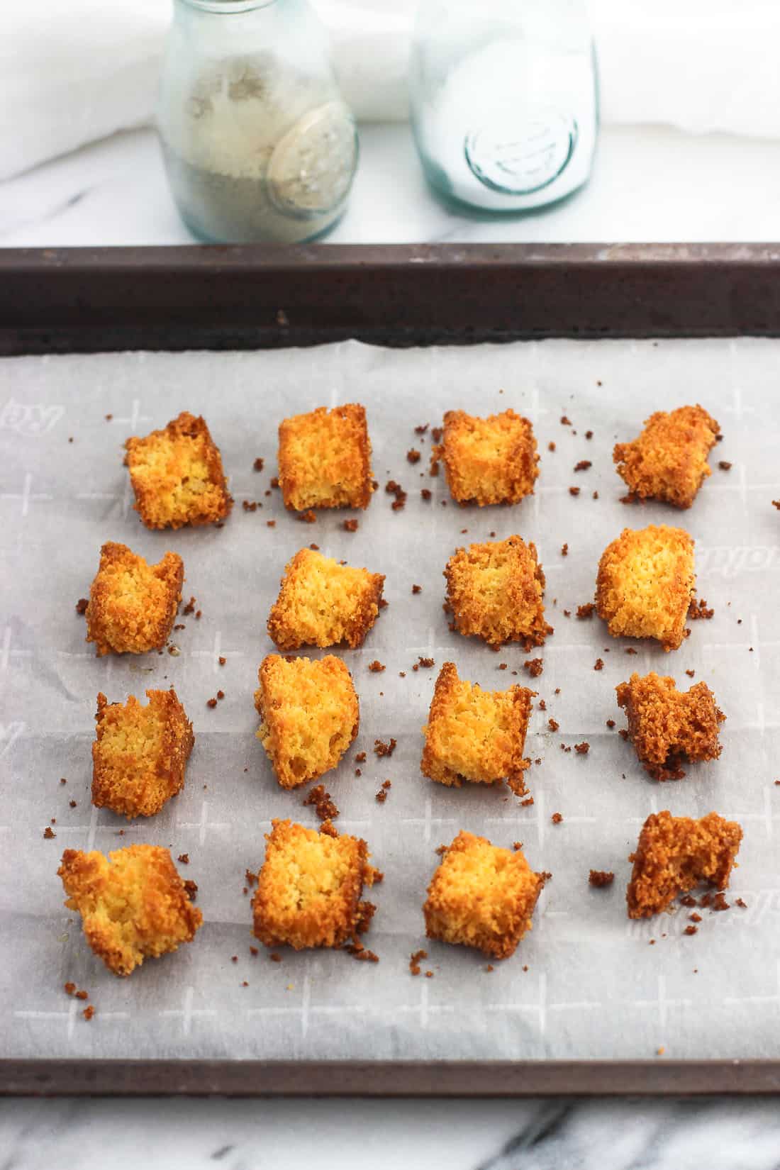 Sixteen cornbread croutons on a parchment-lined baking sheet with salt and pepper shakers in the background