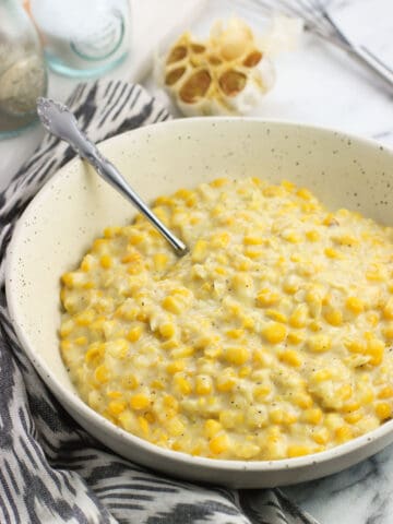 A ceramic serving bowl filled with creamed corn next to a dish towel and a head of roasted garlic