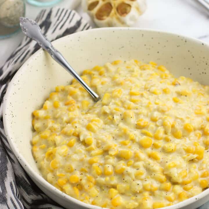 A ceramic serving bowl filled with creamed corn next to a dish towel and a head of roasted garlic