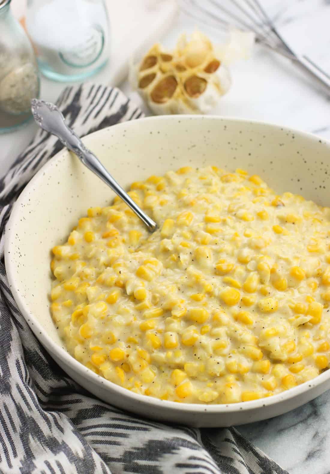 A ceramic serving bowl of creamed corn with a spoon in it next to a dish towel, salt and pepper shakers, and a roasted garlic head