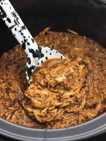 A serving spoon scooped into a slow cooker filled with shredded chicken.