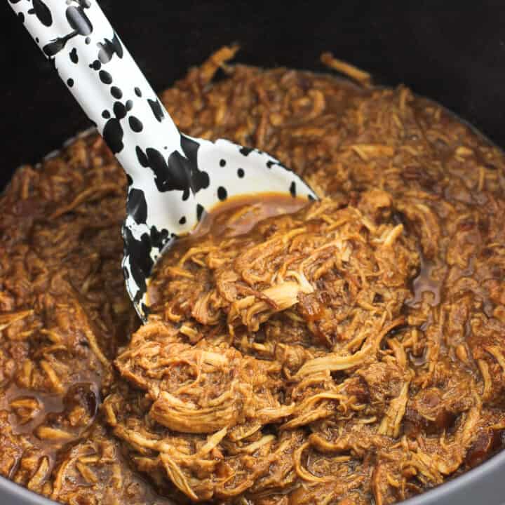 A serving spoon scooped into a slow cooker filled with shredded chicken.