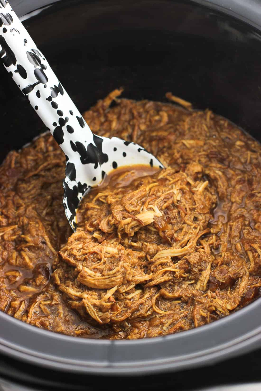 A serving spoon scooped into a slow cooker filled with shredded chicken.